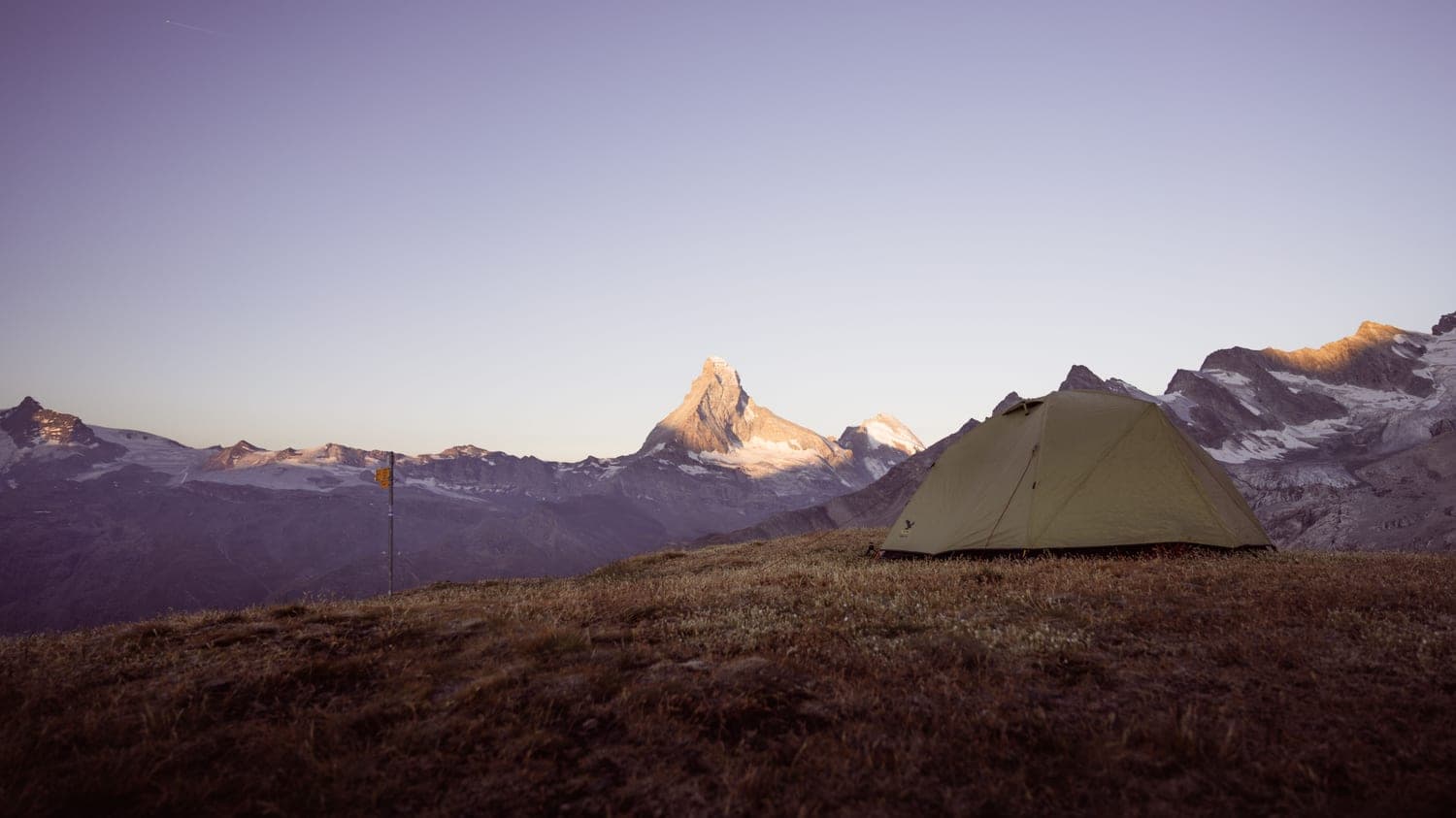 A tent pitched on a grassy ridge below a rocky peak at dusk